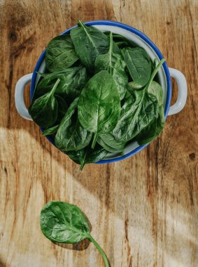 Tahta bir masada taze ıspanak yapraklarıyla dolu beyaz diş minesi tenceresinin yüksek açılı görüntüsü.