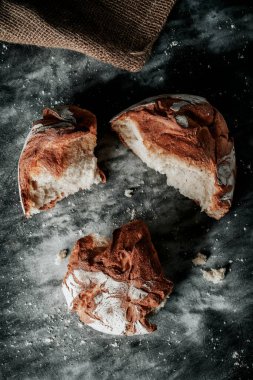 Yuvarlak bir kamut ekmeğinin yüksek açılı görüntüsü bazı parçalar halinde kırılmış, benekli koyu gri bir kumaş yüzeyine yerleştirilmiş, çuval bezinin yanında.