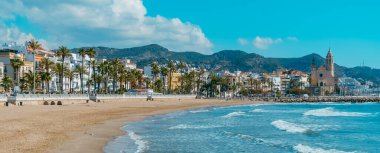 Sitges, Spain - March 3, 2022: A panoramic view of Sitges, Spain, with the Platja de la Fragata beach in the foreground and the Sant Bartomeu i Santa Tecla Church on the right in the background