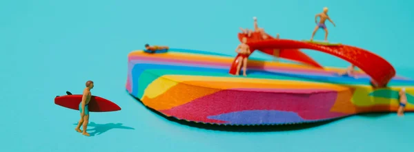 Zbliżenie Miniaturowego Mężczyzny Niosącego Czerwoną Deskę Surfingową Obok Tęczowej Klapki — Zdjęcie stockowe