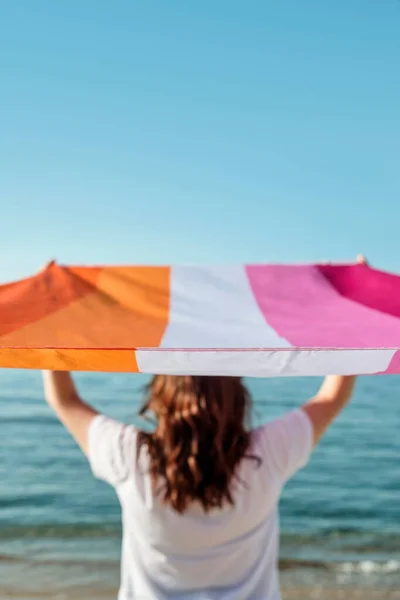 後ろから見た長い黒い髪をした白人女性は海の前で海の上にレズビアンの誇りフラグを立っています — ストック写真