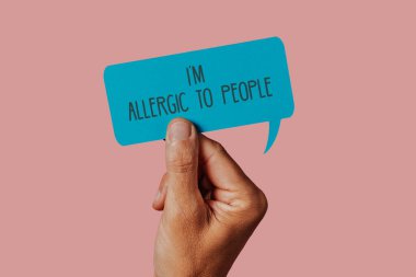 closeup of the hand of a man holding a blue sign in the shape of a speech balloon with the text I am allergic to people written in it, against a pink background clipart