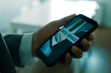 Ofiste elinde akıllı telefonuyla beyaz gömlek ve gri takım elbiseli bir adamı yakından çek. Bu da ekranında, elinde tuttuğu metni okur.