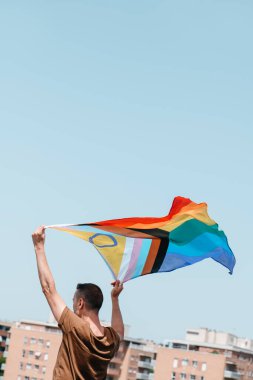 Genç bir adam mavi gökyüzünde, şehirde cinsiyetler arası ilerleme gösteren gurur bayrağı sallıyor.