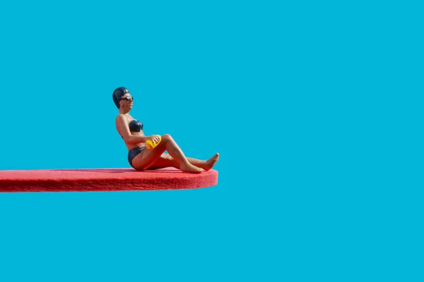 a miniature woman, wearing a black bikingi, a black swimming cap and googles, is sitting on a red diving board, on a blue background