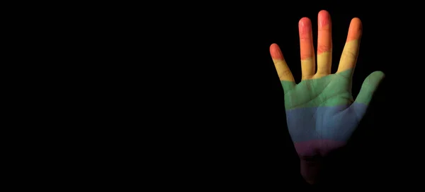 the hand of a person, patterned with the rainbow pride flag, emerges from the black background, with some blank space on the left, in a panoramic format to use as web banner or header