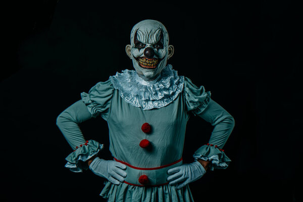a creepy bald evil clown stands against a black background, with hands on hips, and stares at the observer