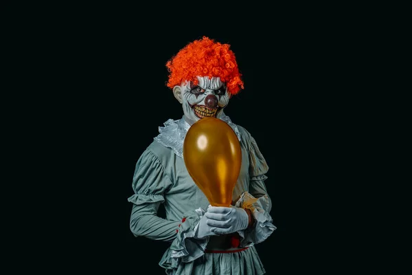 a red-hair creepy evil clown, staring at the observer while is holding a golden balloon in his hands, wearing white gloves, standing on a black background with some blank space around him