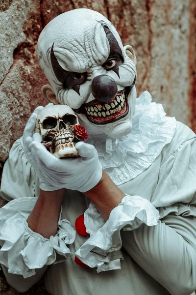 一个毛骨悚然的邪恶小丑 身穿灰色服装 头戴红帽子 手里拿着一个骷髅 站在一座废弃房屋古老生锈的外墙前 — 图库照片
