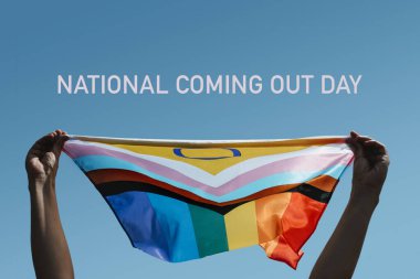 text national coming out day and a young person holding an intersex-inclusive progress pride flag above their head against the blue sky clipart