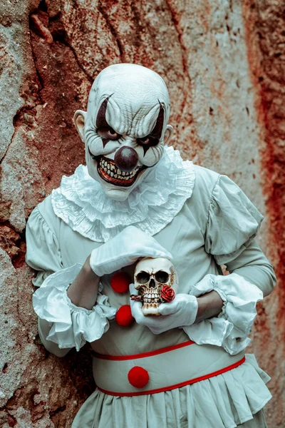 一个毛骨悚然的邪恶小丑的特写镜头 身穿灰色服装 头戴红帽子 头戴白发 手拿着骷髅 靠在一座废弃房屋生锈的外墙上 — 图库照片
