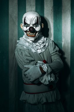 Beyaz yakalı gri bir kostüm giyen tüyler ürpertici kel bir palyaço pis ve eski bir sirk çadırının önünde dikiliyor ve korkunç bir gülümsemeyle gözlemciye bakıyor.