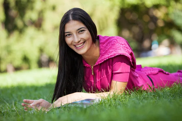 Jovencita alegre tumbada en la hierba en el parque, revista de lectura — Foto de Stock