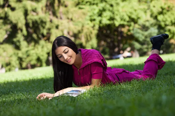 Cheerful young girl lying on grass in park, reading magazine — Stock Photo, Image