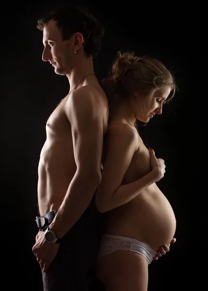 Портрет молодой беременной пары, стоящей спиной к спине на черном фоне — стоковое фото
