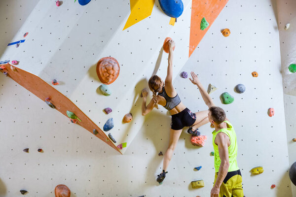 Rock climbers in climbing gym. Young woman climbing bouldering problem (route), man giving her instructions.