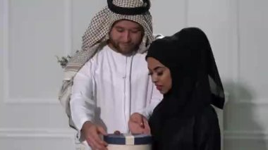 Mutlu Müslüman çift birbirlerine hediyeler veriyor. Romantik Arap adam ve tesettürlü İslami kadın, gri stüdyo arka planında hediyelerini değiş tokuş ediyorlar. Tatilleri birlikte kutluyorlar.