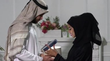 Mutlu Müslüman çift birbirlerine hediyeler veriyor. Romantik Arap adam ve tesettürlü İslami kadın, gri stüdyo arka planında hediyelerini değiş tokuş ediyorlar. Tatilleri birlikte kutluyorlar.