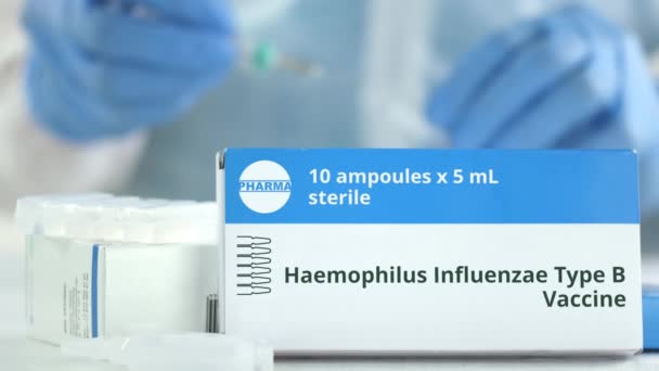 Box mit Haemophilus influenzae Typ B Impfstoff auf dem Tisch gegen verschwommene Laborassistentin. Fiktives pharmazeutisches Logo — Stockvideo