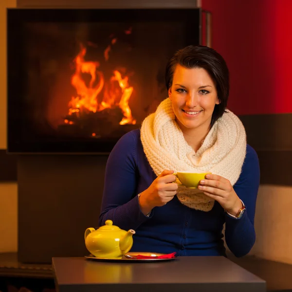 Attraente giovane donna beve il tè in una stanza calda vicino al camino con teiera su un tavolo — Foto Stock