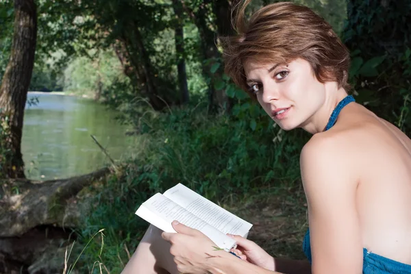 Beautiful young woman sitting by the river and read a book — Stock Photo, Image