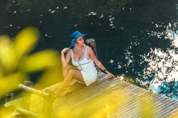 Piękna Blondynka Kapeluszem Głowie Kolorowa Sukienka Siedzi Molo Nad Wodą — Zdjęcie stockowe