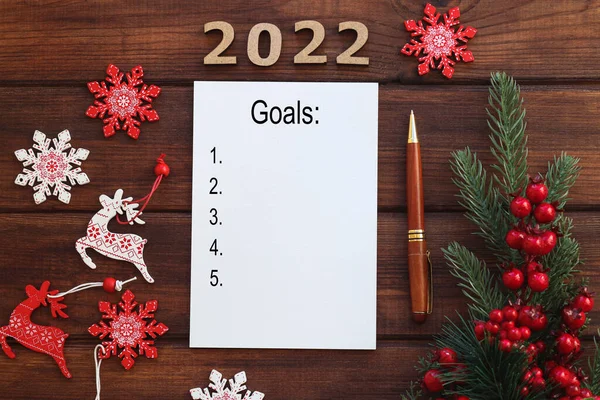 Yeni yıl hedef listesi 2022. Yılın gol ve sonuçlarını yazmak için köknar dalı, kırmızı böğürtlen, kalem ve defter içeren şenlikli bir arka plan. Üst Manzara. Tatil ve motivasyon kavramı