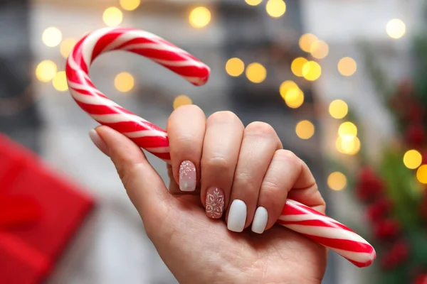 Vrouwen hand met gel polish manicure witte kleur en met sneeuwvlokken ornament, met snoep stok tegen feestelijke kerst achtergrond. Selectieve focus. Idee van de winter manicure — Stockfoto