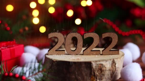 Schnee fällt auf die Holzzahlen 2022. Festlicher Hintergrund für das neue Jahr mit Weihnachtsschmuck, Lichtern, Tannenzweigen und Bokeh. Nahaufnahme, selektiver Fokus — Stockvideo