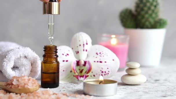 L'huile sérique coule du compte-gouttes. Set de massage à l'huile essentielle, sel d'himalaya rose, bougie parfumée, fleur d'orchidée, pierres zen et serviette. Concept beauté et spa — Video