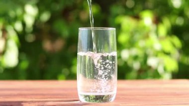 Temiz su, yemyeşil yapraklara karşı masanın üzerindeki bardağa dökülüyor. Detoks ve yaz ferahlatıcı içecek konsepti.