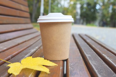Empty cup of coffee and yellow leaf on bench against background of blurred autumn park. Copy space or mock up.