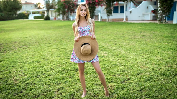 young thin blonde girl stands barefoot smilr on a green lawn in a summer dress and holds a straw hat