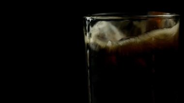İki parça buz, siyah arka planda siyah soda ile bir bardağa düşer.