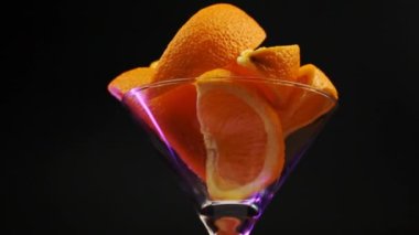 Martini bardağındaki portakallar ortada mor bir tonda siyah bir zemin üzerinde dönüyorlar.
