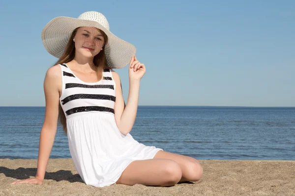 Vrouw in witte jurk en hoed zittend op het strand — Stockfoto