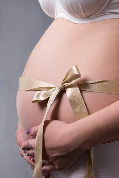 Bliska ciąży brzuch z Dar złote wstążki na szary — Zdjęcie stockowe