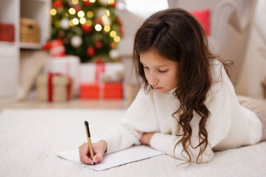 Noel ağacı ve hediyelerle süslenmiş oturma odasında Noel Baba 'ya mektup yazan küçük tatlı bir kızın portresi.