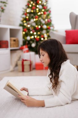 Noel ağacıyla süslü odada kitap okuyan tatlı bir kızın portresi.