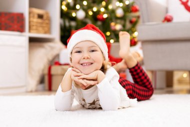 Mutlu küçük kız evdeki Noel ağacının yanındaki halıda yatıyor.