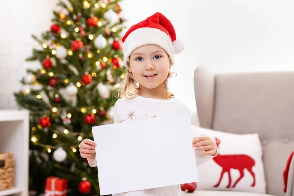 女の子ホームでクリスマスツリーの近くに空白の紙を保持 — ストック写真