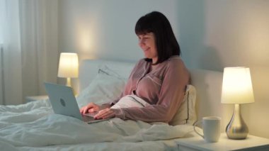 Genç bir kadın arkadaşlarıyla video görüşmesi yapıyor. Laptop kullanıyor. Geceleri yatakta oturuyor.