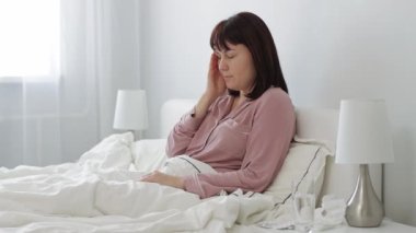 Baş ağrısı olan genç hasta bir kadın yatağında oturmuş, suyla birlikte hap alıyor.