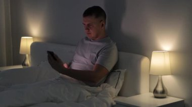 Genç adam yatağında oturuyor ve gece karanlık yatak odasında video görüşmesi için akıllı telefon kullanıyor.