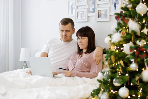 クリスマスとオンラインショッピングのコンセプト 幸せなカップルがベッドに座って クリスマスツリーで装飾された寝室でオンラインで何かを購入 — ストック写真
