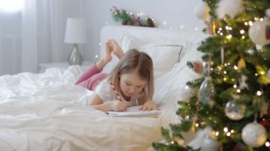Noel ağacıyla süslenmiş yatak odasında Noel Baba 'ya mektup yazan tatlı küçük kız.