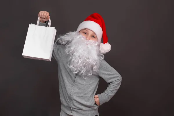 Santa boy with beard and hat holds a white paper eco-friendly gift bag. copy space — Stock Photo, Image