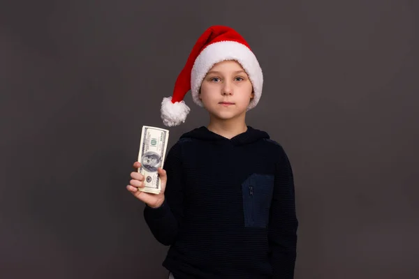 The boy is holding dollars in Santas helper hat in his hand. on a gray background. mock up — Stock Photo, Image