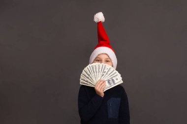 Dolar hayranı bir çocuk yüzünü kapattı. Gri arka planda Noel Baba şapkalı bir çocuk. Boşluğu kopyala