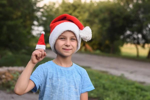 Funny smiling boy in Santas helper hat. in the summer on the street. Against the background of green foliage. — Stock Photo, Image